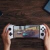 control-8bitdo-ultimate-mobile-gaming Person holds a white handheld gaming console playing a racing game on a wooden table, with a phone and a tablet nearby.