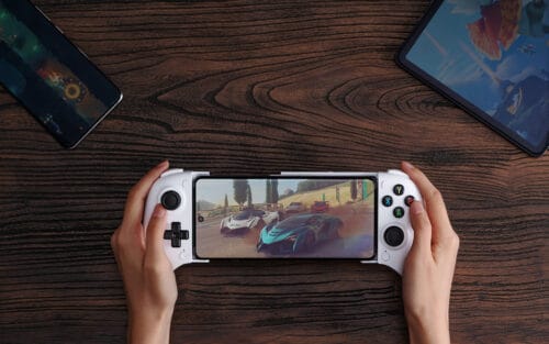 control-8bitdo-ultimate-mobile-gaming Person holds a white handheld gaming console playing a racing game on a wooden table, with a phone and a tablet nearby.