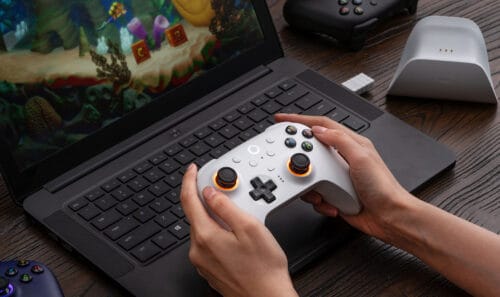 02-l Person holding a white game controller over a laptop keyboard with a colorful game on the screen behind it.