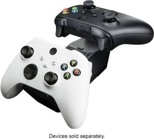 07e2f3fc-db52-4f60-8b43-6bb78da0a172 Two Xbox-style wireless controllers (white and black) resting on a charging dock, with the note 'Devices sold separately' shown below.