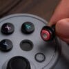 61HikWYNk2L._SL1500_ Close-up of a hand inserting a wireless earbud into a circular game controller with colorful X and A buttons.