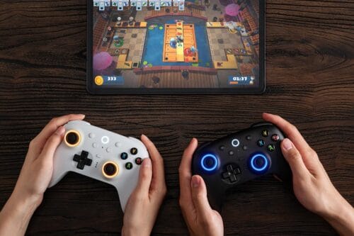 Version 1.0.0 Two people hold white and black game controllers while playing a board-style video game on a tablet screen above a wooden table.
