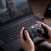 Version 1.0.0 Person holding an Xbox-style controller while gaming on a laptop on a wooden desk, screen showing gameplay.