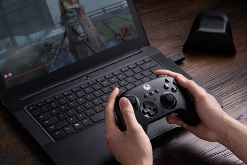 Version 1.0.0 Person holding an Xbox-style controller while gaming on a laptop on a wooden desk, screen showing gameplay.