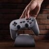 71R1hIXsTWL._SL1500_ Close-up of a hand pressing a gray retro-style game controller resting on a curved dock against a brick wall.