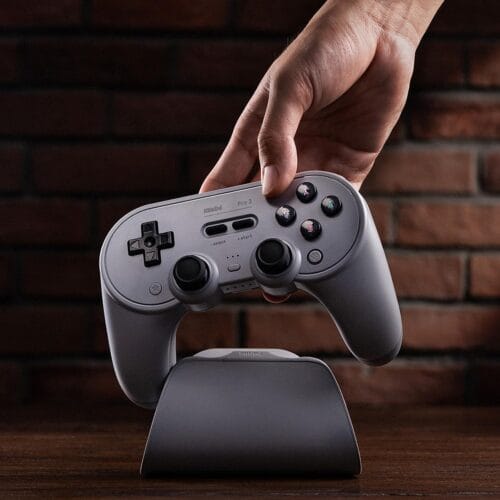 71R1hIXsTWL._SL1500_ Close-up of a hand pressing a gray retro-style game controller resting on a curved dock against a brick wall.