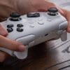 Version 1.0.0 Close-up of hands holding a gray game controller on a wooden surface, showing the joysticks and buttons.