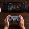 71ugO+7W5KL._SL1500_ Hands hold a gray retro-style game controller in front of a black handheld gaming device displaying an action game on a wooden table.