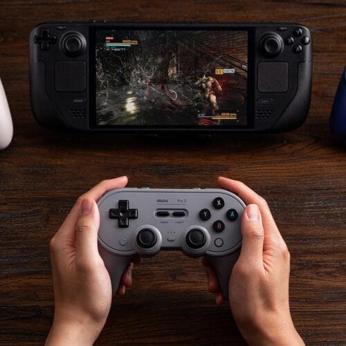 71ugO+7W5KL._SL1500_ Hands hold a gray retro-style game controller in front of a black handheld gaming device displaying an action game on a wooden table.