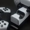 next-accessories-playstation-NXPS5CHRG2-54184593-1-1024x677 (1) Two white wireless game controllers sit on a white charging dock with a central handset and battery charging contacts visible.