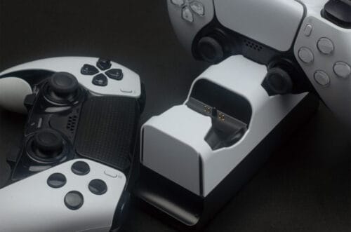 next-accessories-playstation-NXPS5CHRG2-54184593-1-1024x677 (1) Two white wireless game controllers sit on a white charging dock with a central handset and battery charging contacts visible.