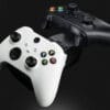 next-accessories-xbox-NXXCHRG2-54190022-1-1024x683 White Xbox-style controller and a black controller on a charging dock against a dark background, angled diagonally left to right in the frame.