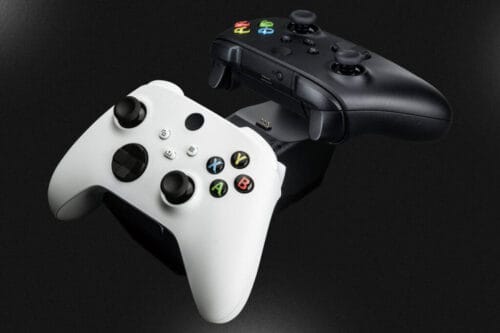 next-accessories-xbox-NXXCHRG2-54190022-1-1024x683 White Xbox-style controller and a black controller on a charging dock against a dark background, angled diagonally left to right in the frame.