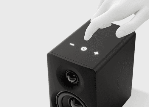 Hand tapping the touch controls (−, power, +) on a small black speaker.