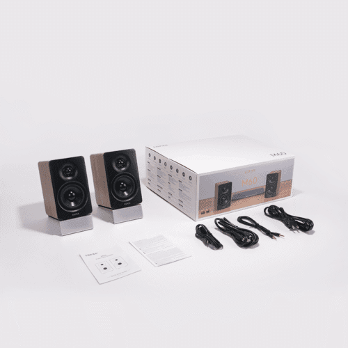 Two wooden bookshelf speakers with black fronts and silver bases, beside a white M60 box with cables and manuals nearby.
